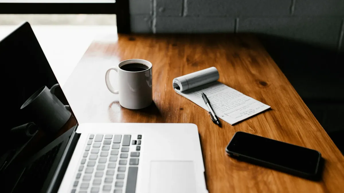Laptop, coffee mug and notebook on a desk beside a bright window