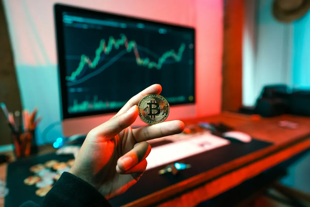 Hand holding a physical Bitcoin in front of a trading monitor
