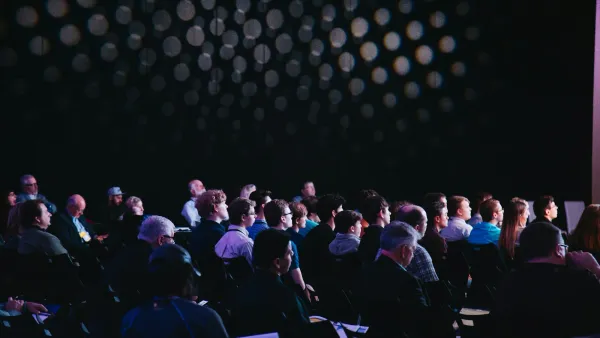 Large audience seated in a dark conference hall facing the stage